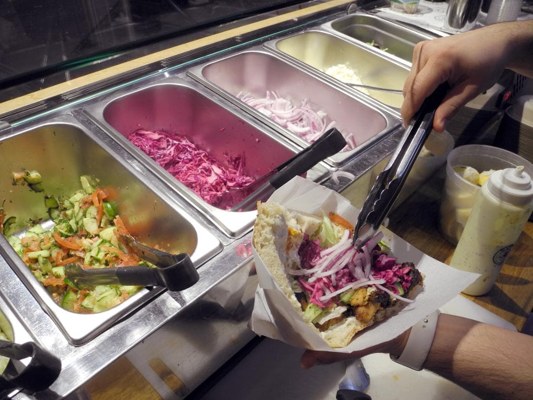 Hinter der Theke: Edelstahlbehälter mit Gurken-Tomaten-Salat, Rotkohl und Zwiebeln, daneben eine Soßenflasche; eine Hand legt mit der Zange Zwiebeln auf.